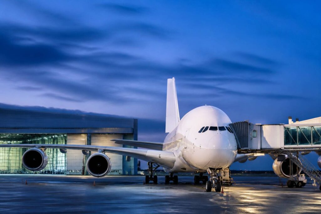 Avion de ligne gros-porteur blanc garé sur un tarmac au crépuscule avec passerelle et hangar de maintenance